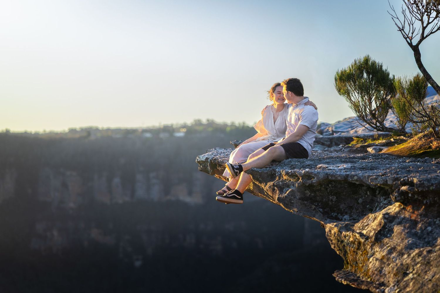 Silhouetted couple sits on rocky cliff edge at Lincoln's Rock, Wentworth Falls overlooking a dramatic landscape at sunset.