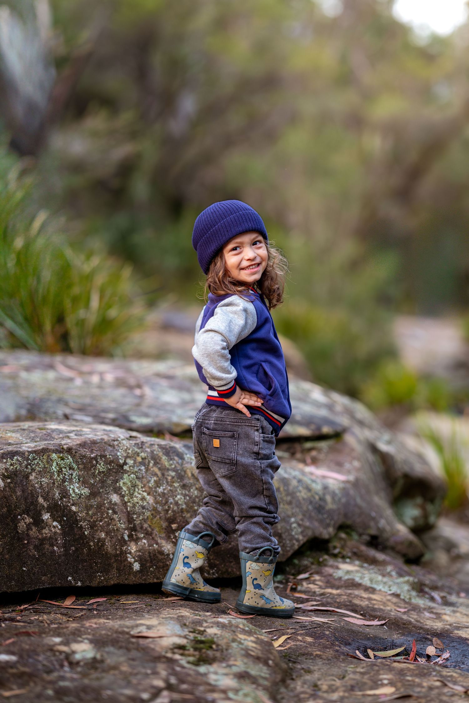 A smiling toddler in a blue beanie, navy vest, and rain boots poses on rocks in a lush forest setting at Maya Sanctuary.