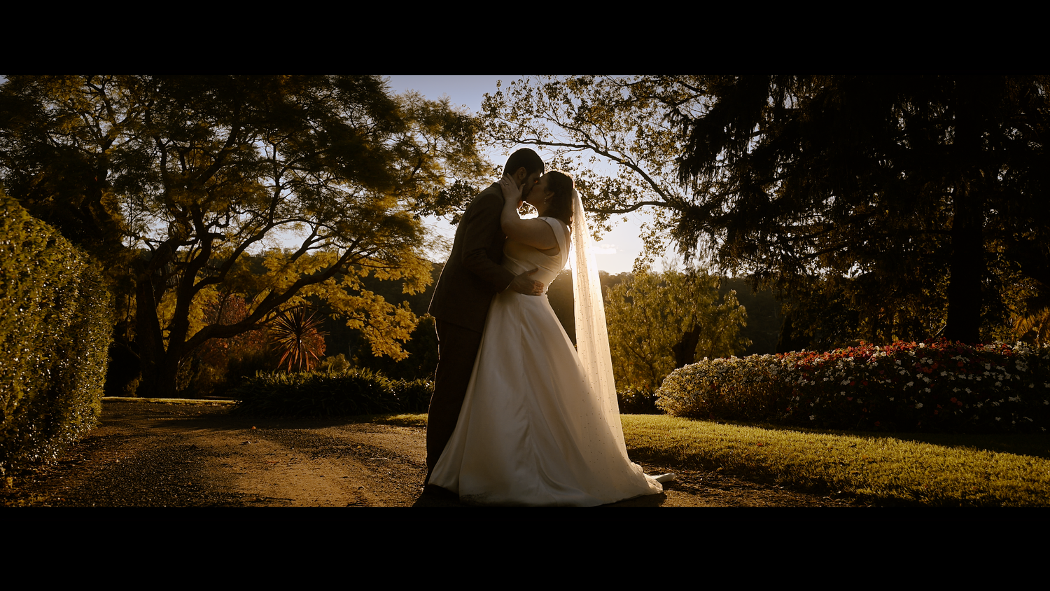 A couple shares a romantic kiss in a sunlit garden at Glenleigh Estate, the bride in a white gown and veil, surrounded by lush greenery.