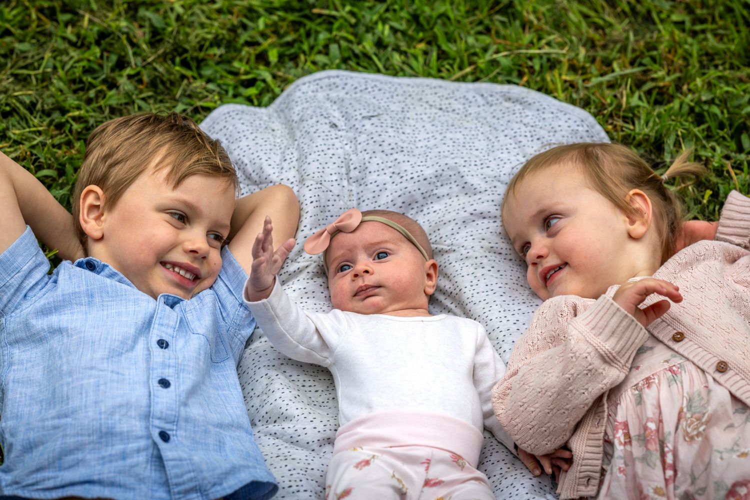 Three young siblings lying on a blanket in grass at Tench Reserve, Nepean River. Two toddlers smiling at a newborn baby between them.