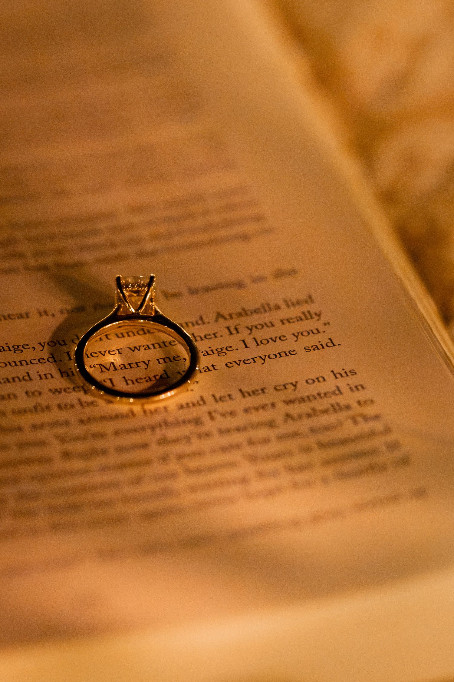 Gold diamond engagement ring resting on open book page with romantic marriage proposal text in warm light.