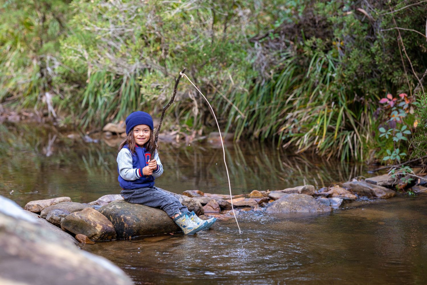 Young child sitting on rocks in a stream, holding a fishing stick, surrounded by lush green forest at Maya Sanctuary.