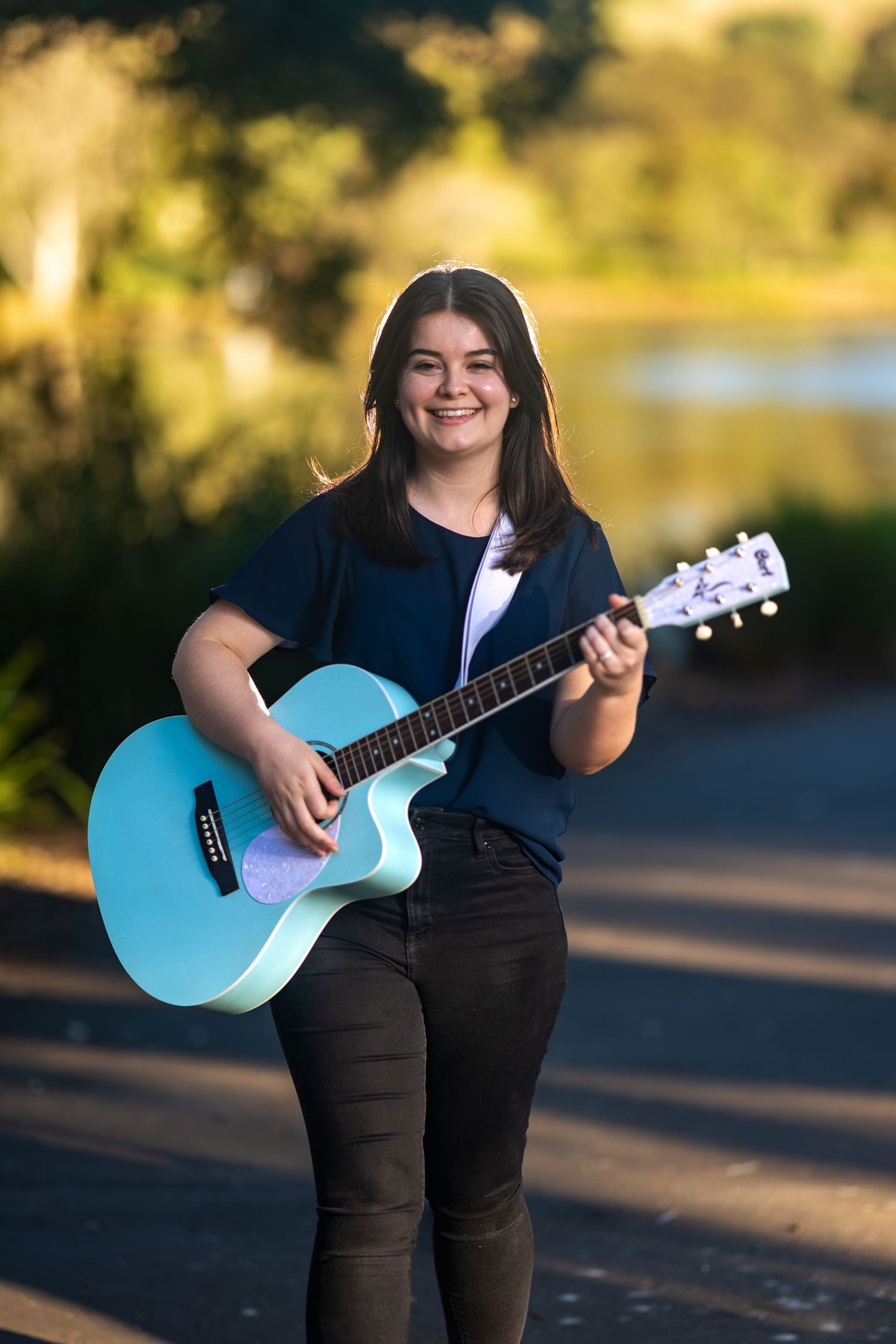 Smiling young woman playing a light blue acoustic guitar outdoors on a road with blurred green trees behind her.