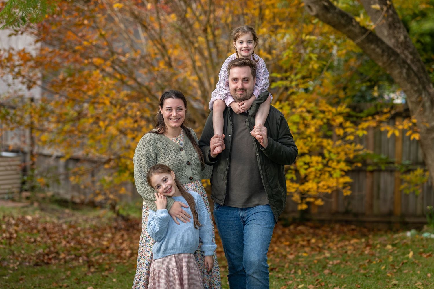 Family photo in Winmalee, Blue Mountains posing outdoors in autumn park with colourful fall foliage in background.