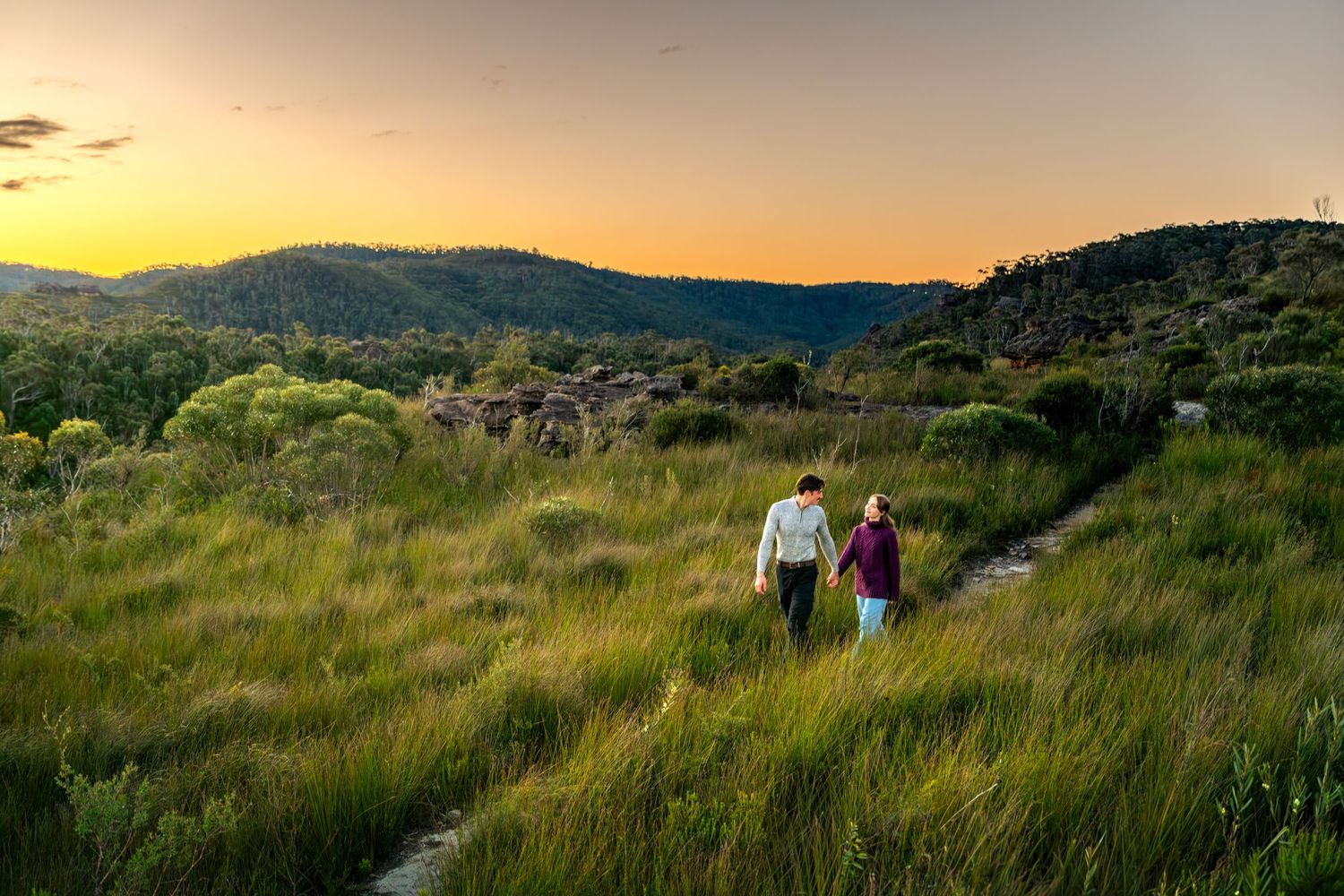 Couple walking hand in hand along a grassy trail at sunset with lush green hills in the background at Walls Lookout in the Blue Mountains.