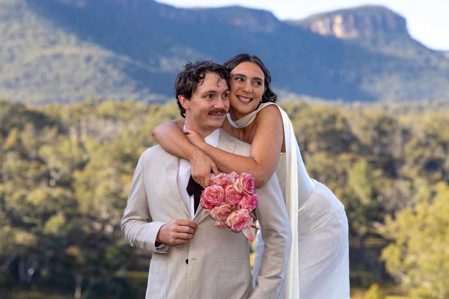 Happy couple embracing outdoors on wedding day, bride holding pink bouquet with mountain backdrop at Maya Sanctuary.