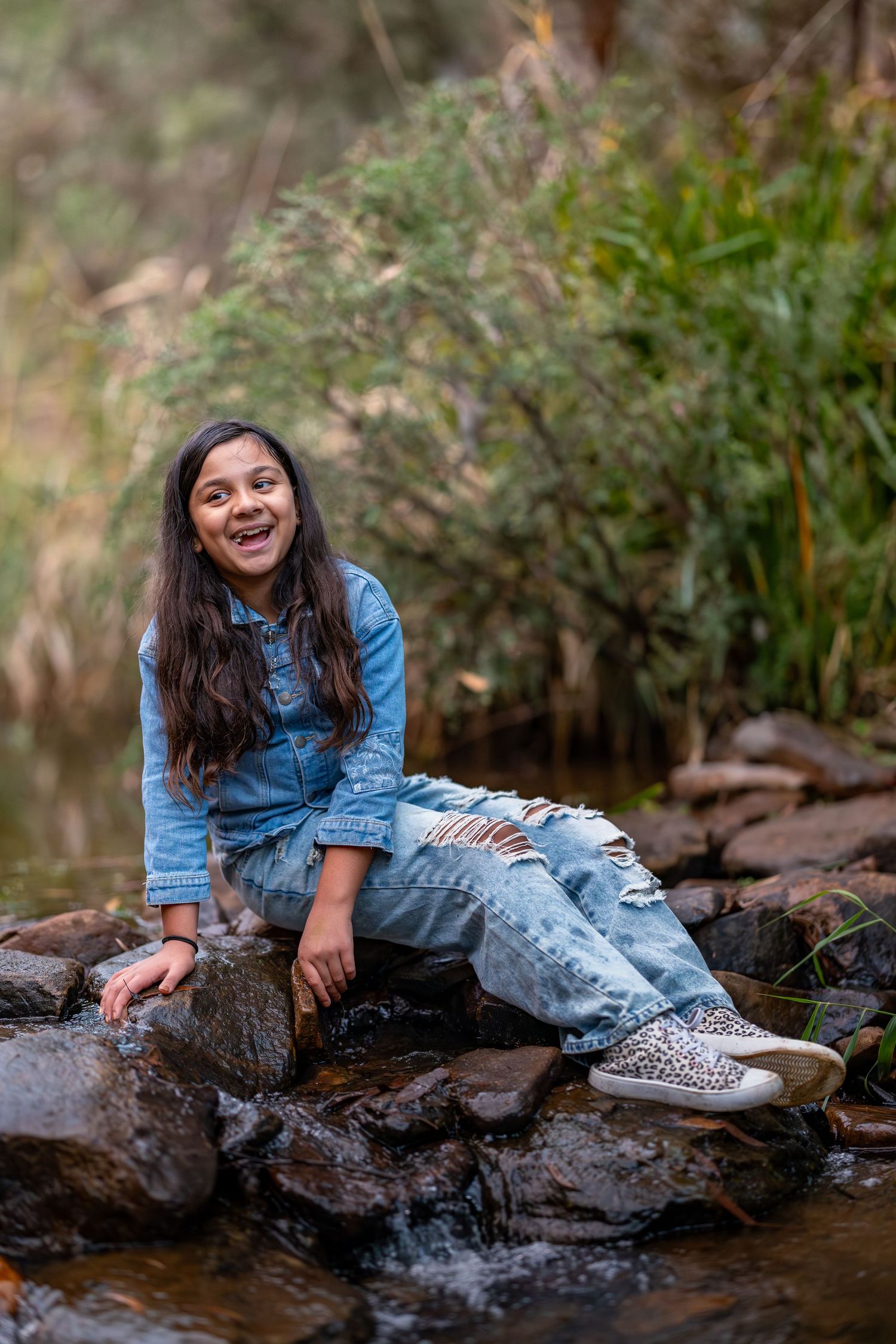 A smiling girl in a denim jacket and ripped jeans sits on rocks by a stream in a lush green forest at Maya Sanctuary.