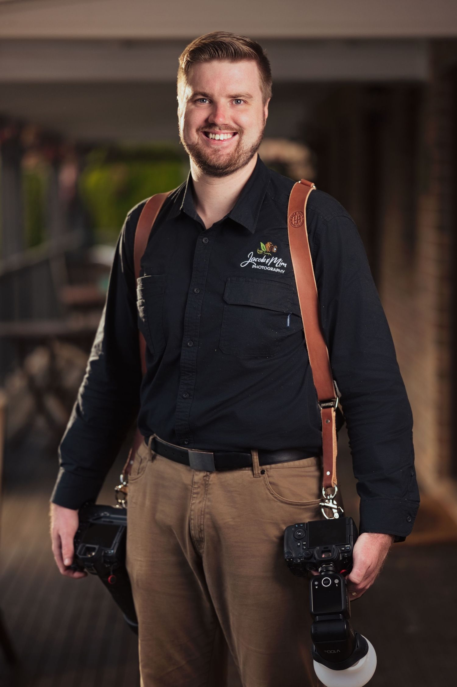 Smiling male photographer in black shirt holding two cameras with brown leather straps in an indoor setting.