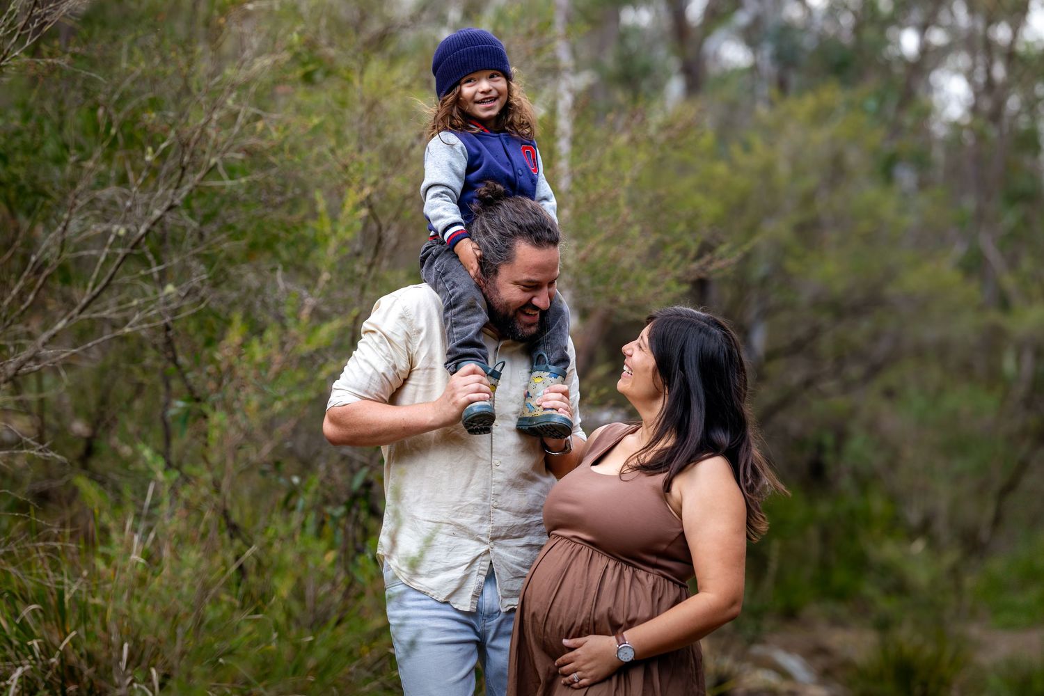 Pregnant woman and man with toddler on shoulders smiling outdoors in nature for a maternity family photo session at Maya Sanctuary.