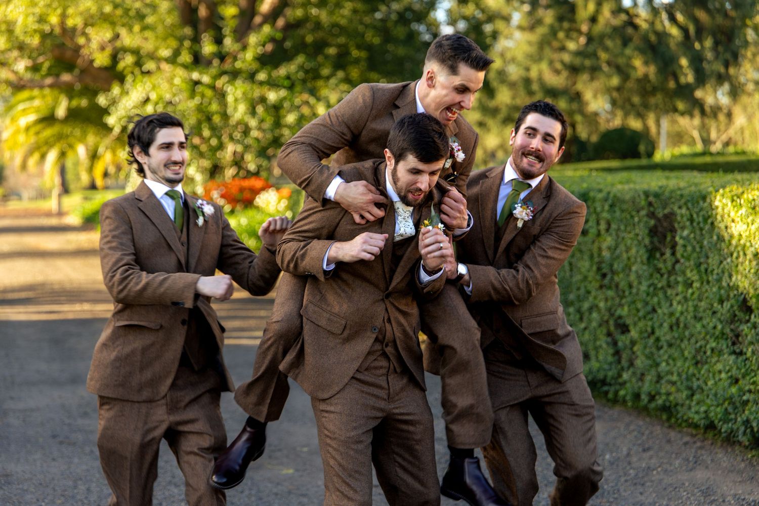 Groomsmen in brown suits laughing and joking together outdoors at a wedding celebration in a garden setting at Glenleigh Estate.