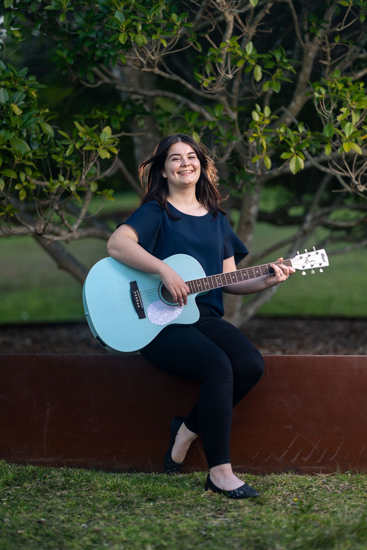 Smiling young woman sitting outdoors playing a light blue acoustic guitar, wearing black outfit with trees in background.