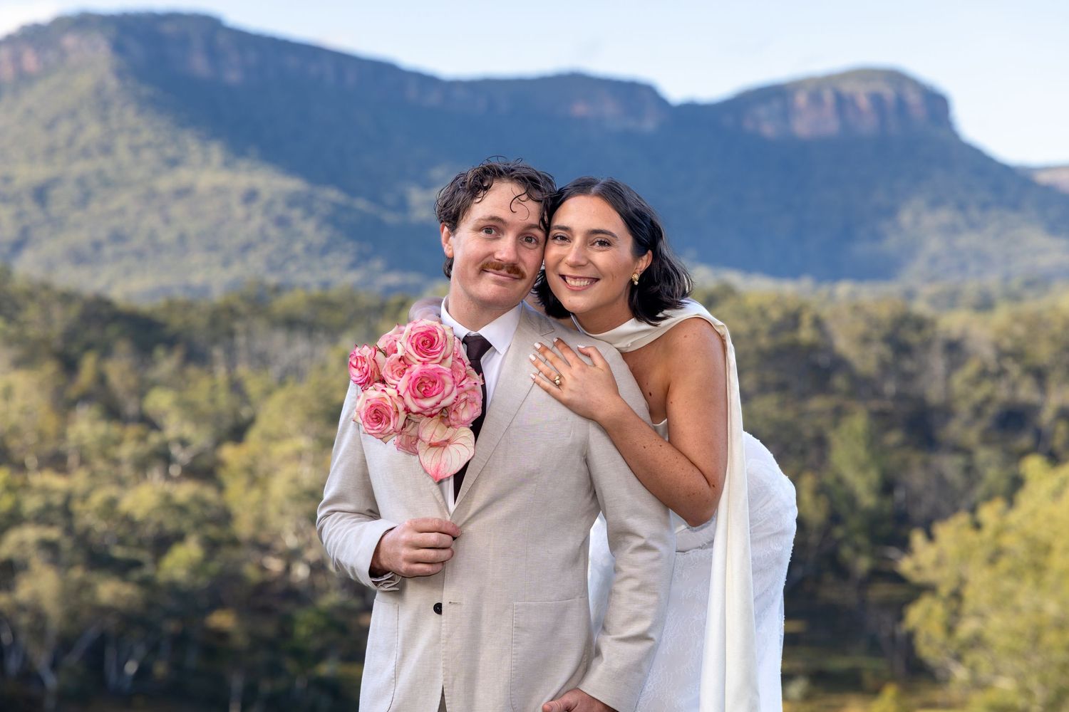 Bride and groom smiling with pink roses, mountains and lush greenery in background at outdoor wedding at Maya Sanctuary.