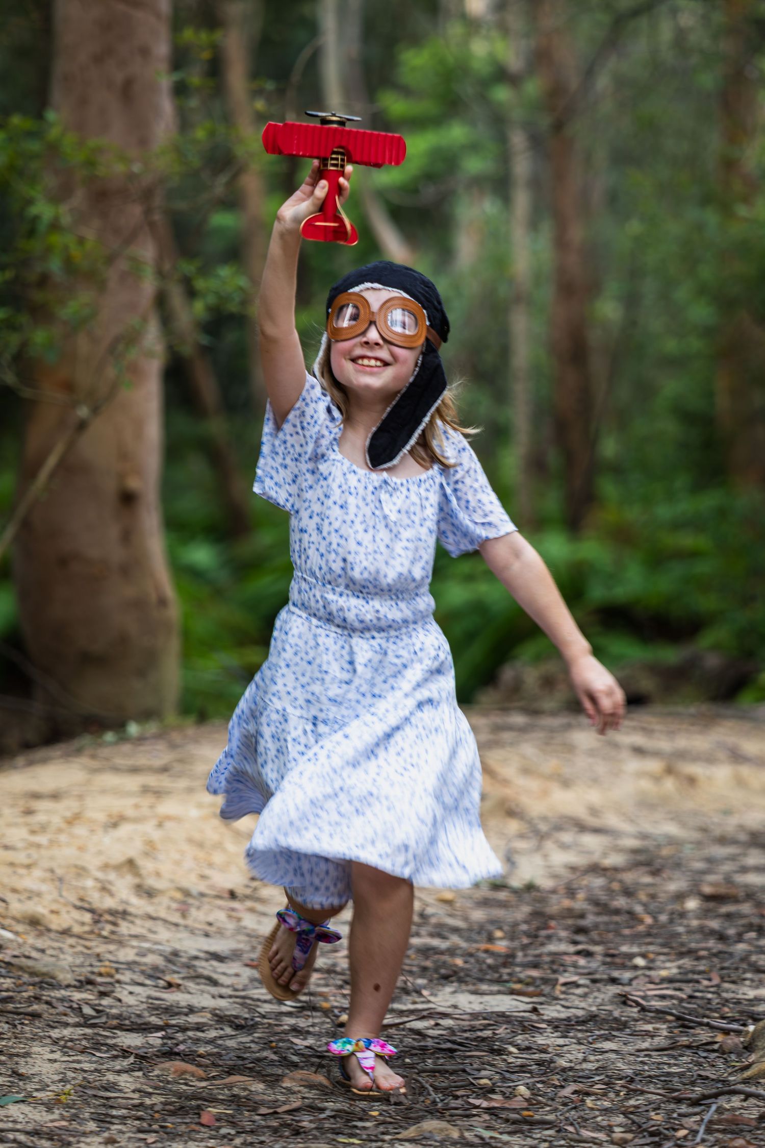 Girl in blue dress and aviator goggles runs through forest holding red toy airplane aloft, smiling joyfully.