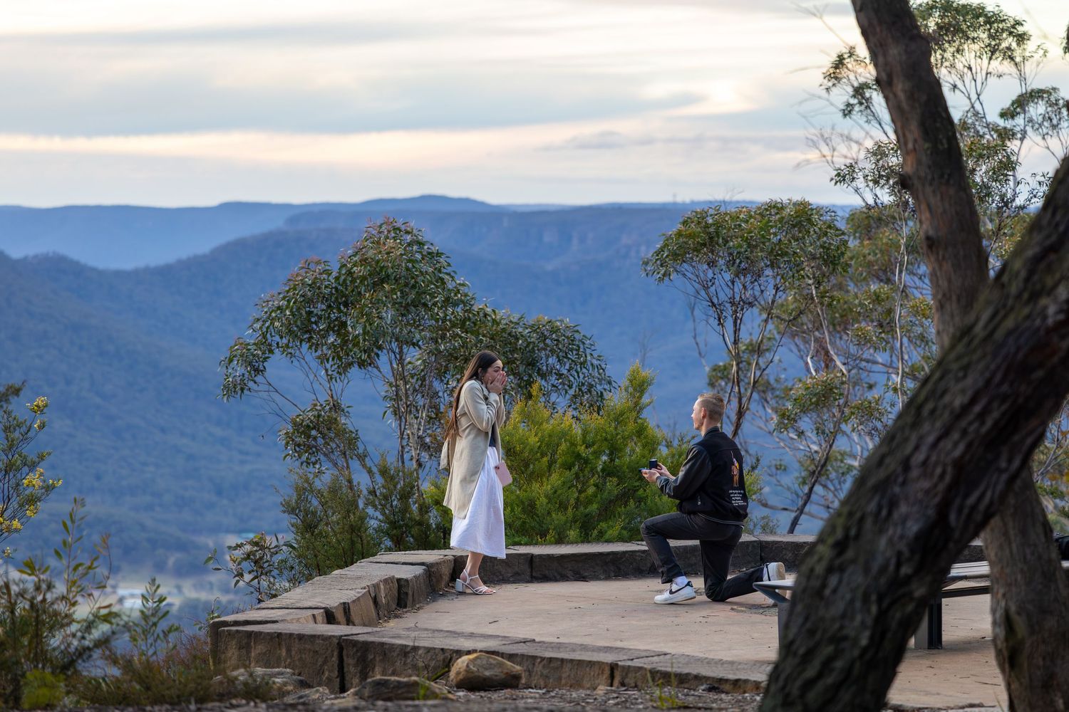 Man proposes to woman on wooden deck overlooking the Blue Mountains valley with lush trees and misty peaks at Mount Blackheath Lookout.