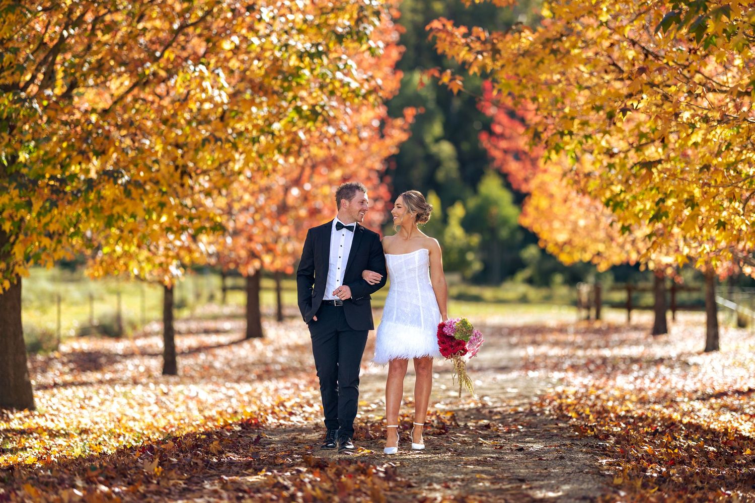 Bride and groom walk hand-in-hand along a leaf-covered path surrounded by vibrant autumn trees at Lawson Park, Hartley Vale.
