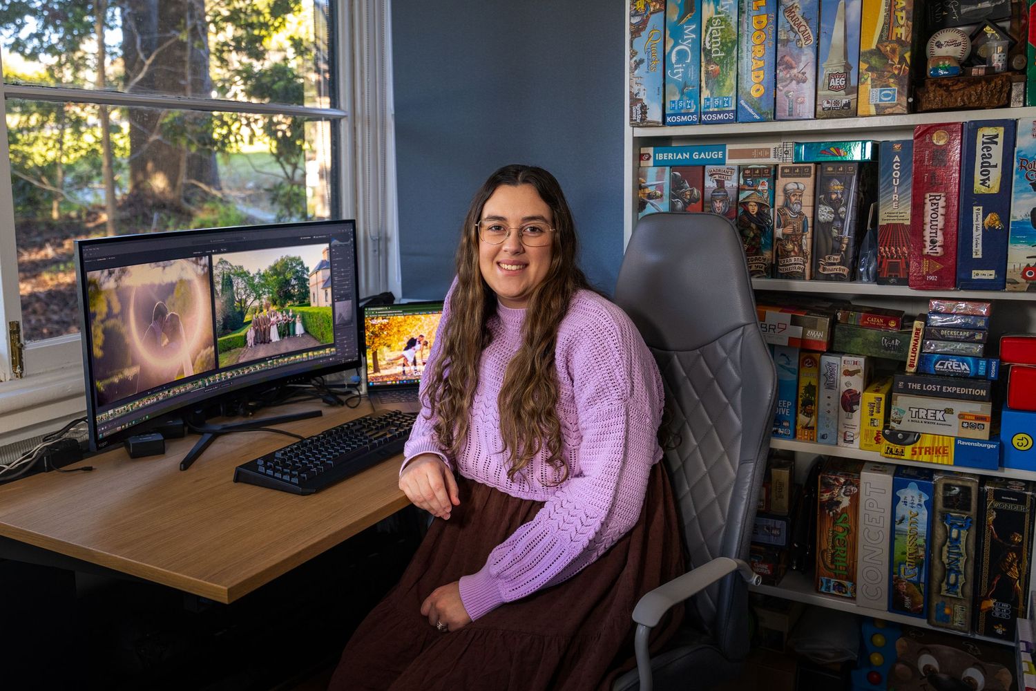 Woman smiling at desk with dual monitors and board game collection shelves in home office setup.