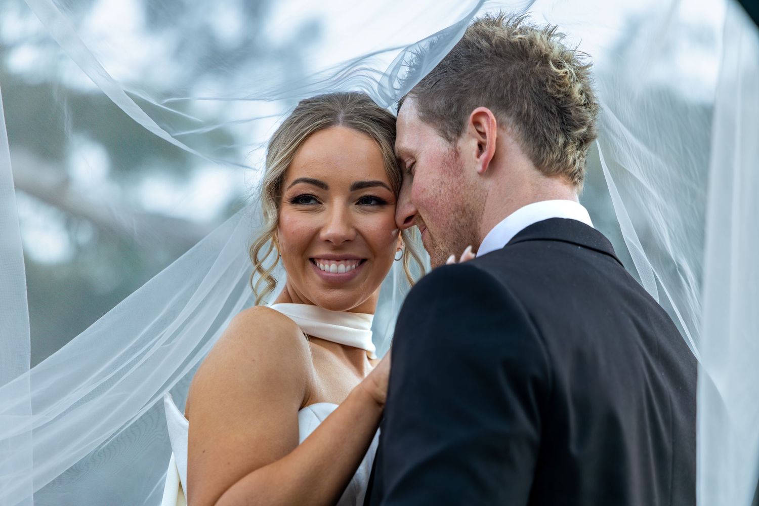 Bride smiling under veil with groom in black suit sharing intimate moment at outdoor wedding at Lawson Park, Hartley Vale.