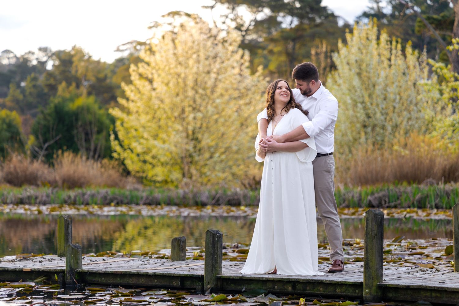 Couple embracing on a wooden dock at Blackheath, NSW, surrounded by autumn foliage and a serene pond, woman in white dress.