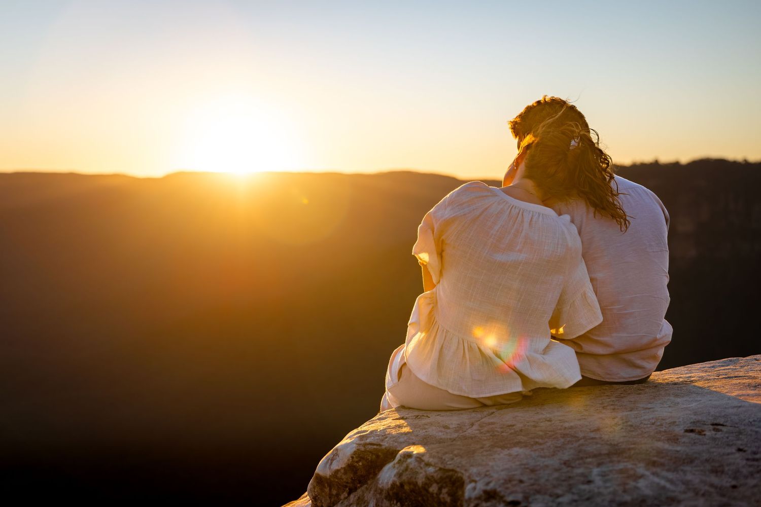 Two silhouettes sit together on a cliff edge watching a golden sunset over mountains in the distance.