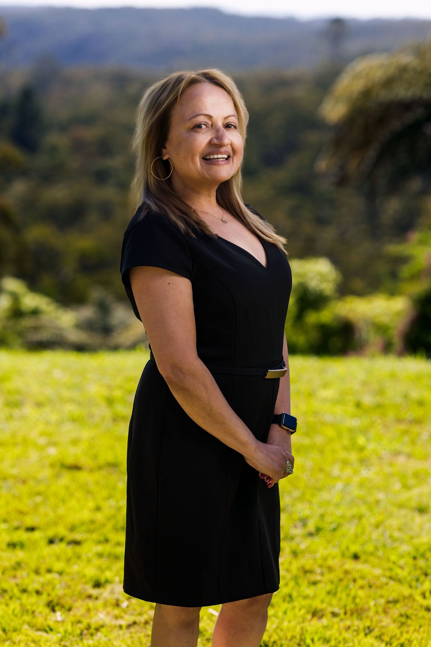 Smiling woman in black dress standing on green grass with lush tropical trees in background.