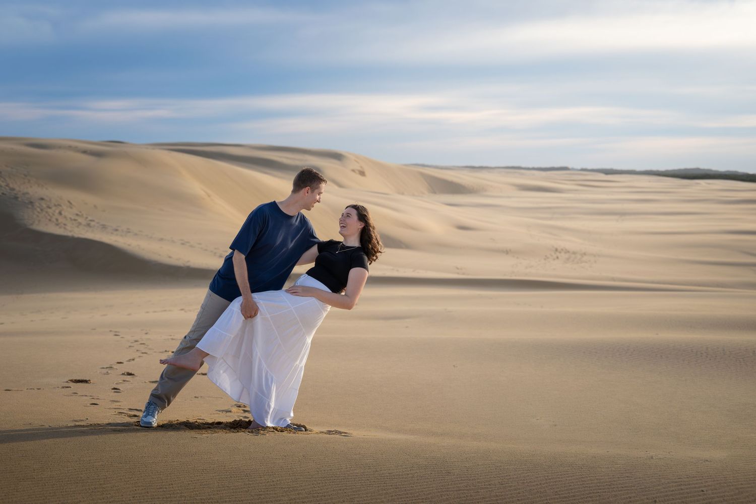 Couple dancing romantically on sandy desert dunes under a blue sky, woman in white skirt and man in blue shirt.