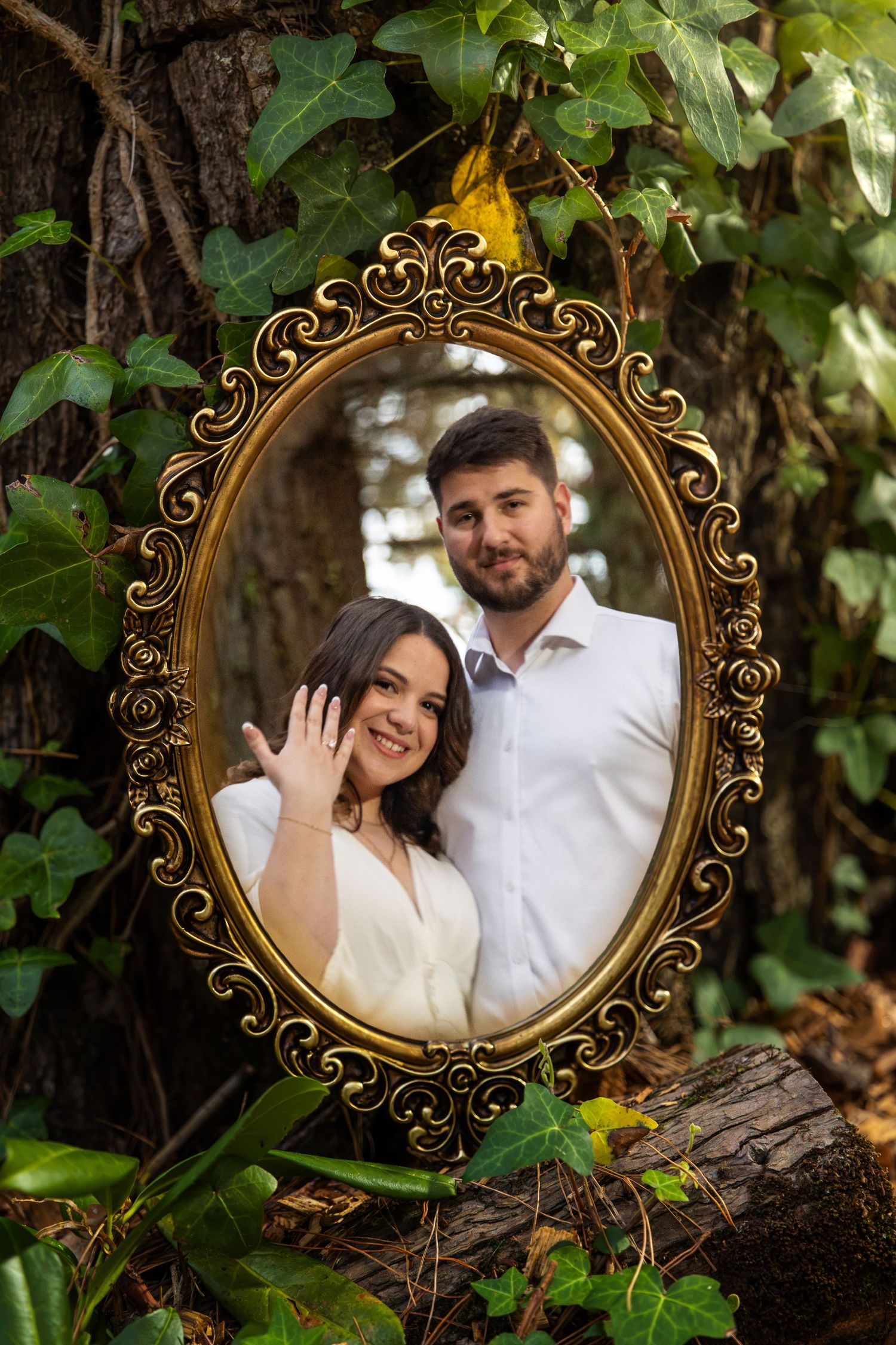 Couple smiling in ornate gold oval mirror surrounded by ivy, woman showing engagement ring in forest setting.