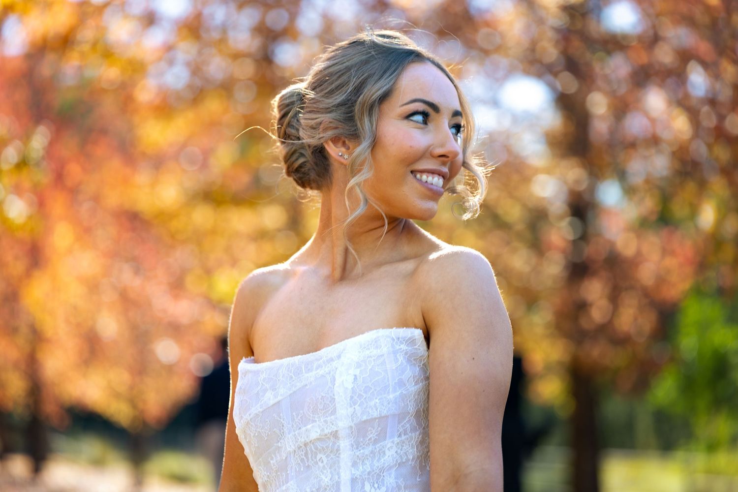Woman in white strapless dress smiling outdoors with vibrant autumn foliage in warm orange and red tones at Lawson Park, Hartley Vale.