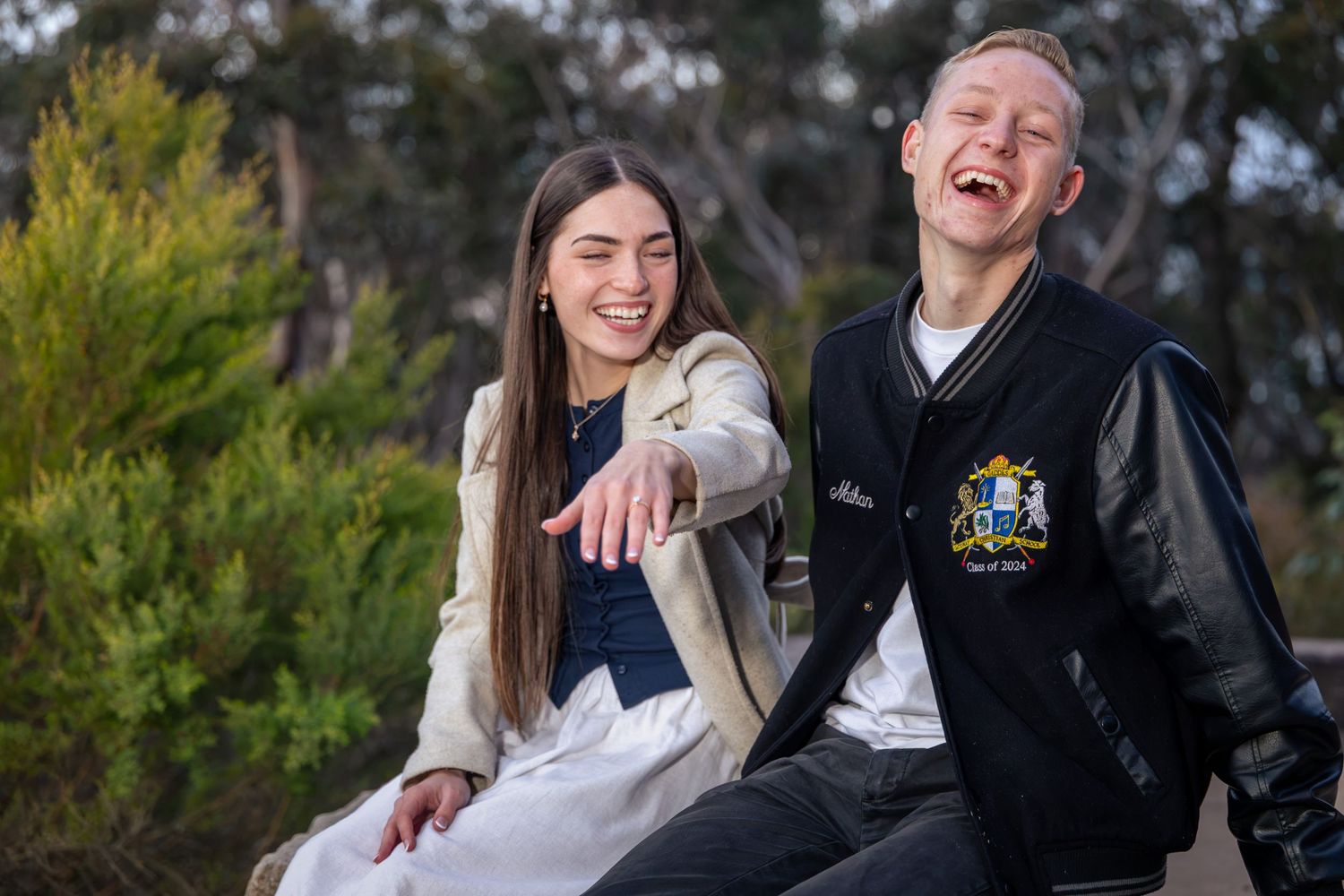 Two smiling teenagers sitting outdoors, girl in white dress and boy in varsity jacket, laughing together in natural setting.