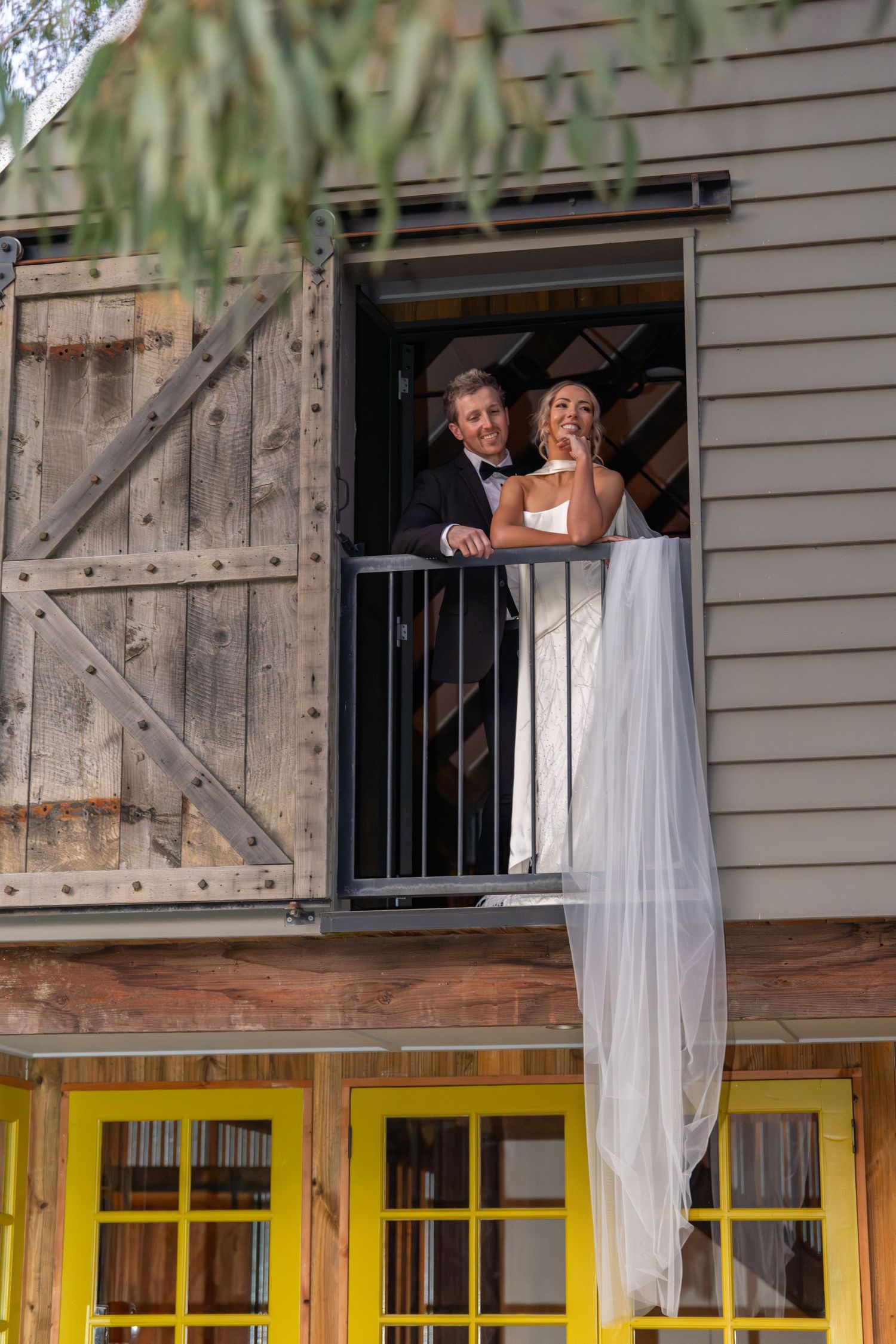 Bride and groom stand on rustic barn balcony, her veil flowing down, smiling joyfully on their wedding day at Lawson Park, Hartley Vale.