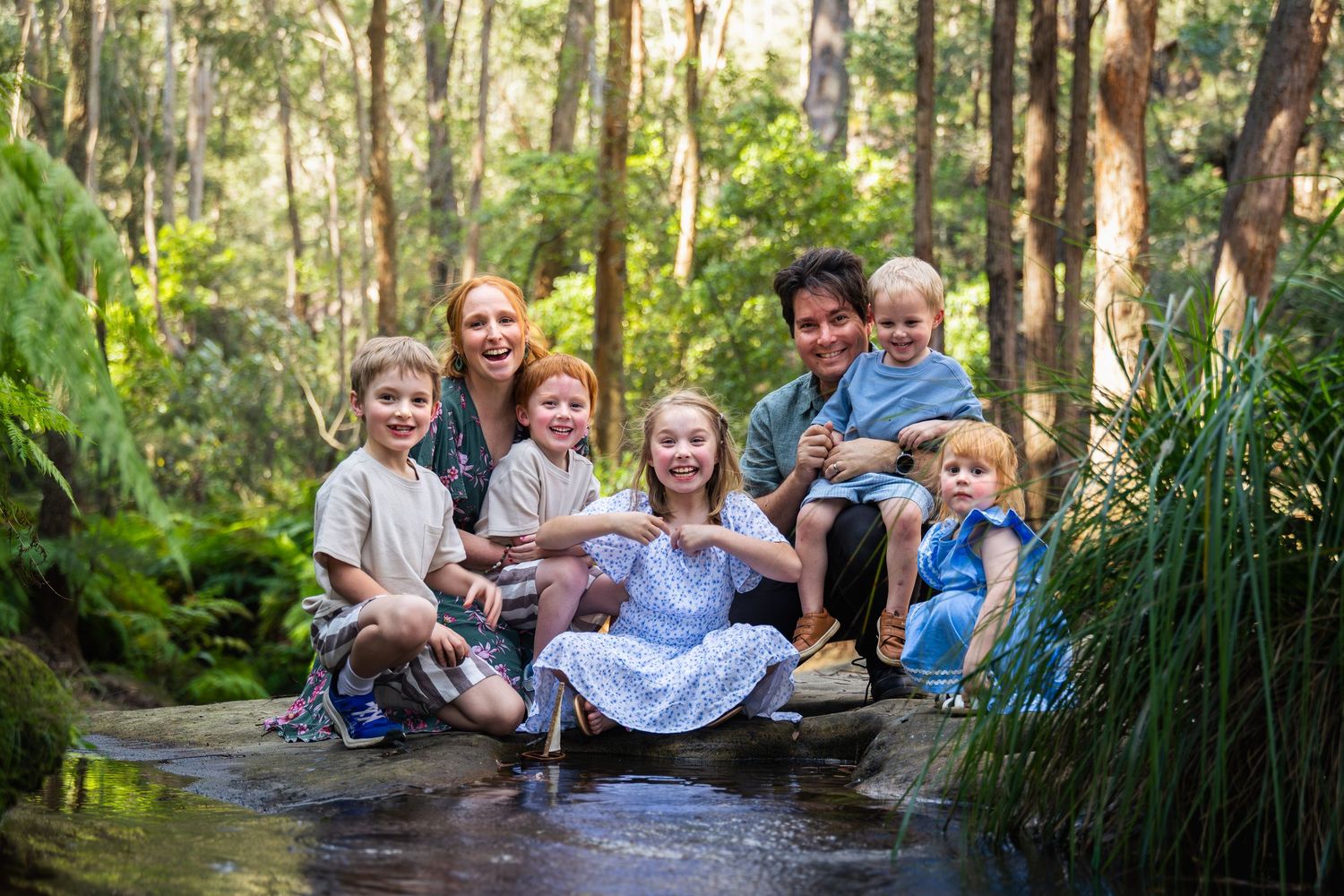A joyful family with five children poses together in a lush green forest stream, smiling for a portrait.