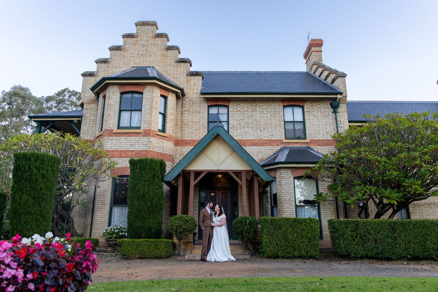 Bride and groom embrace before a grand historic brick manor at with gabled roofline and lush gardens at Glenleigh Estate.