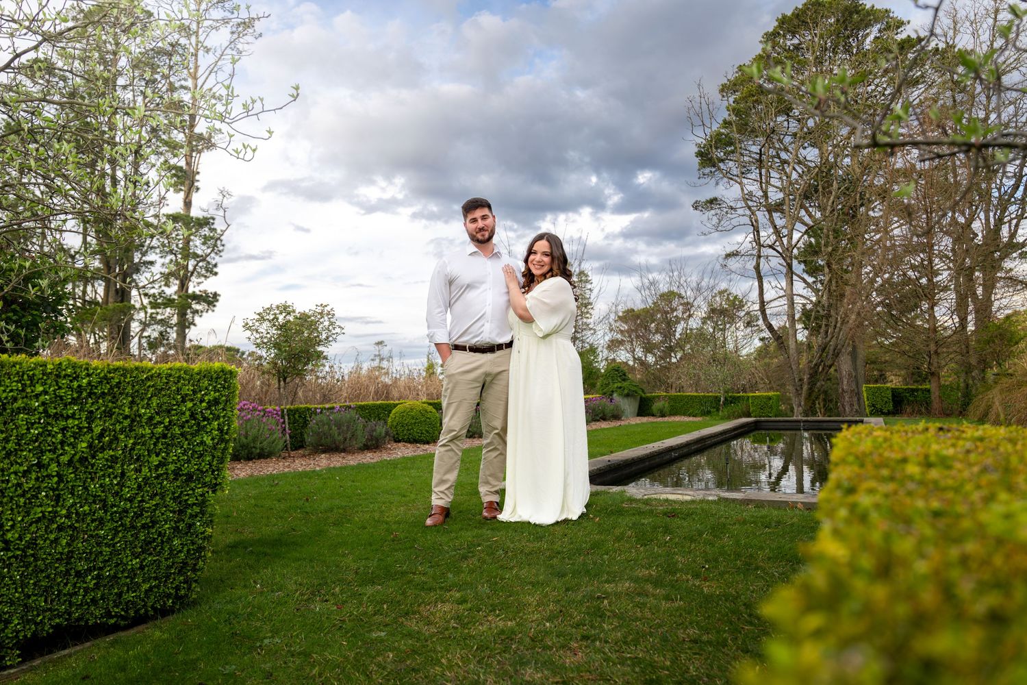 Couple posing in lush green garden with hedges, pond, and colorful flowers under cloudy sky in Blackheath, Blue Mountains.