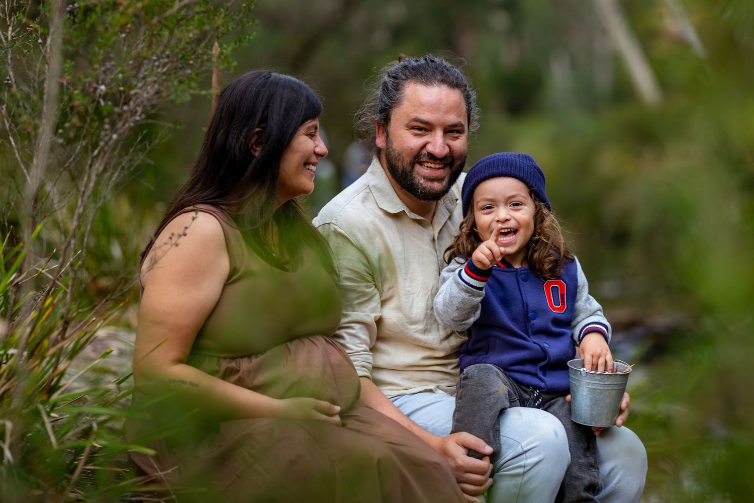 A happy family of three smiling outdoors in lush greenery, with a toddler in a blue hat holding a cup at Maya Sanctuary.