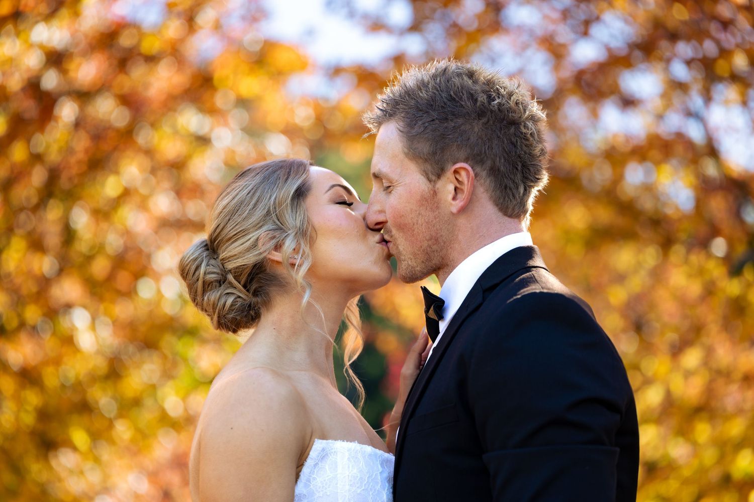 Bride and groom sharing a romantic kiss outdoors with vibrant autumn foliage in shades of orange and gold at Lawson Park, Hartley Vale.