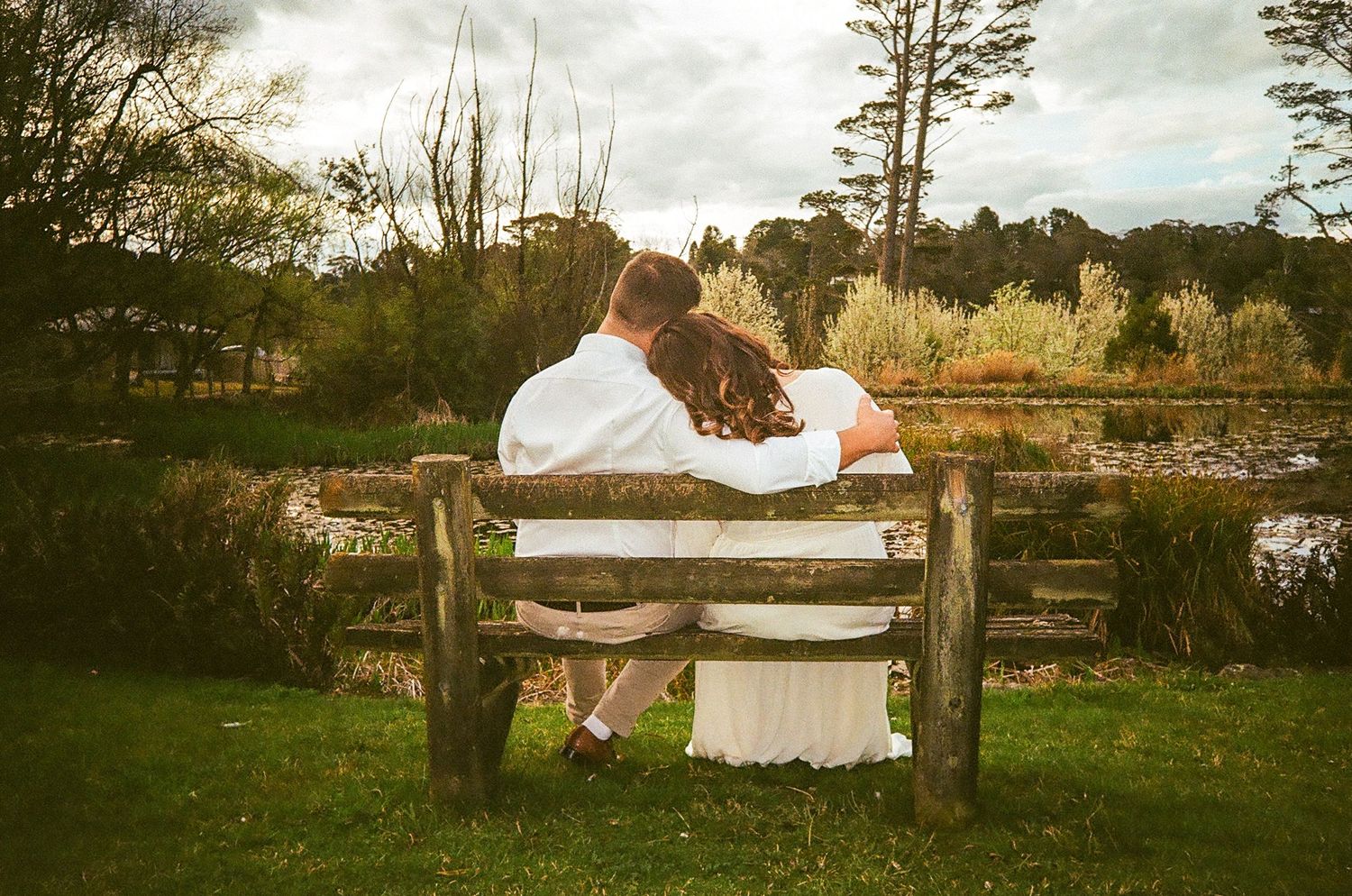 Couple sitting together on a rustic wooden bench by a serene pond, viewed from behind in wedding attire.