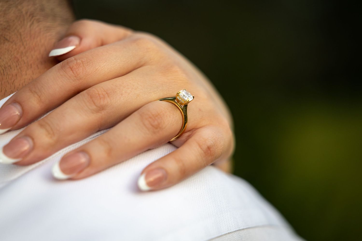 Close-up of a woman's hand with French manicure wearing a gold diamond engagement ring resting on a man's shoulder.