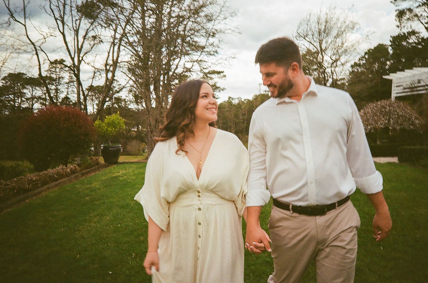 Happy couple walking hand in hand through a lush green garden, smiling at each other on a sunny day.
