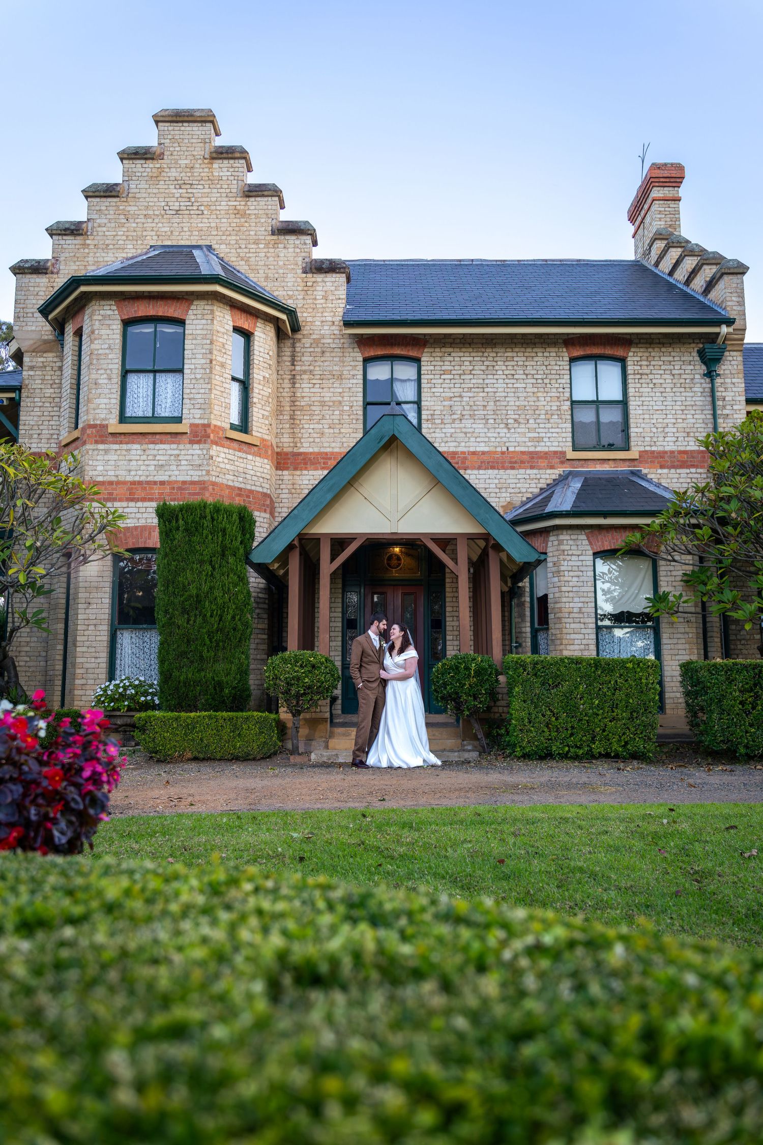 Bride and groom embrace before a grand brick Victorian mansion at Glenleigh Estate with lush gardens at dusk.