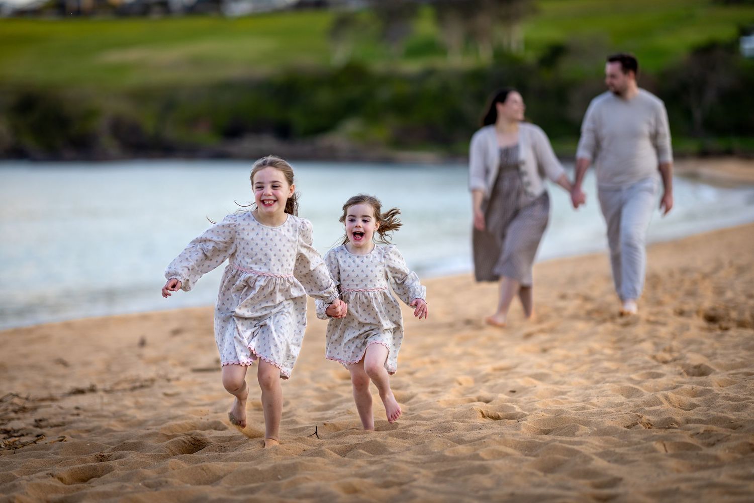 A family at Easts Beach, Kiama runs and plays together on a sandy beach at sunset wearing coordinated white outfits.