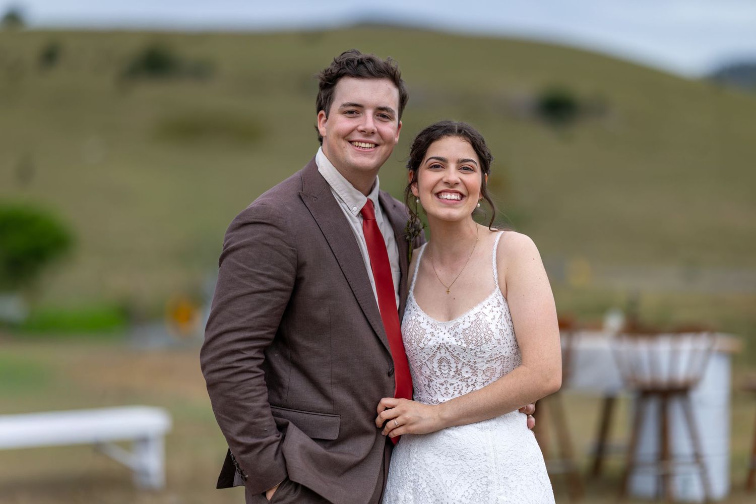 A couple in wedding attire poses together in front of a scenic rural landscape with rolling hills.