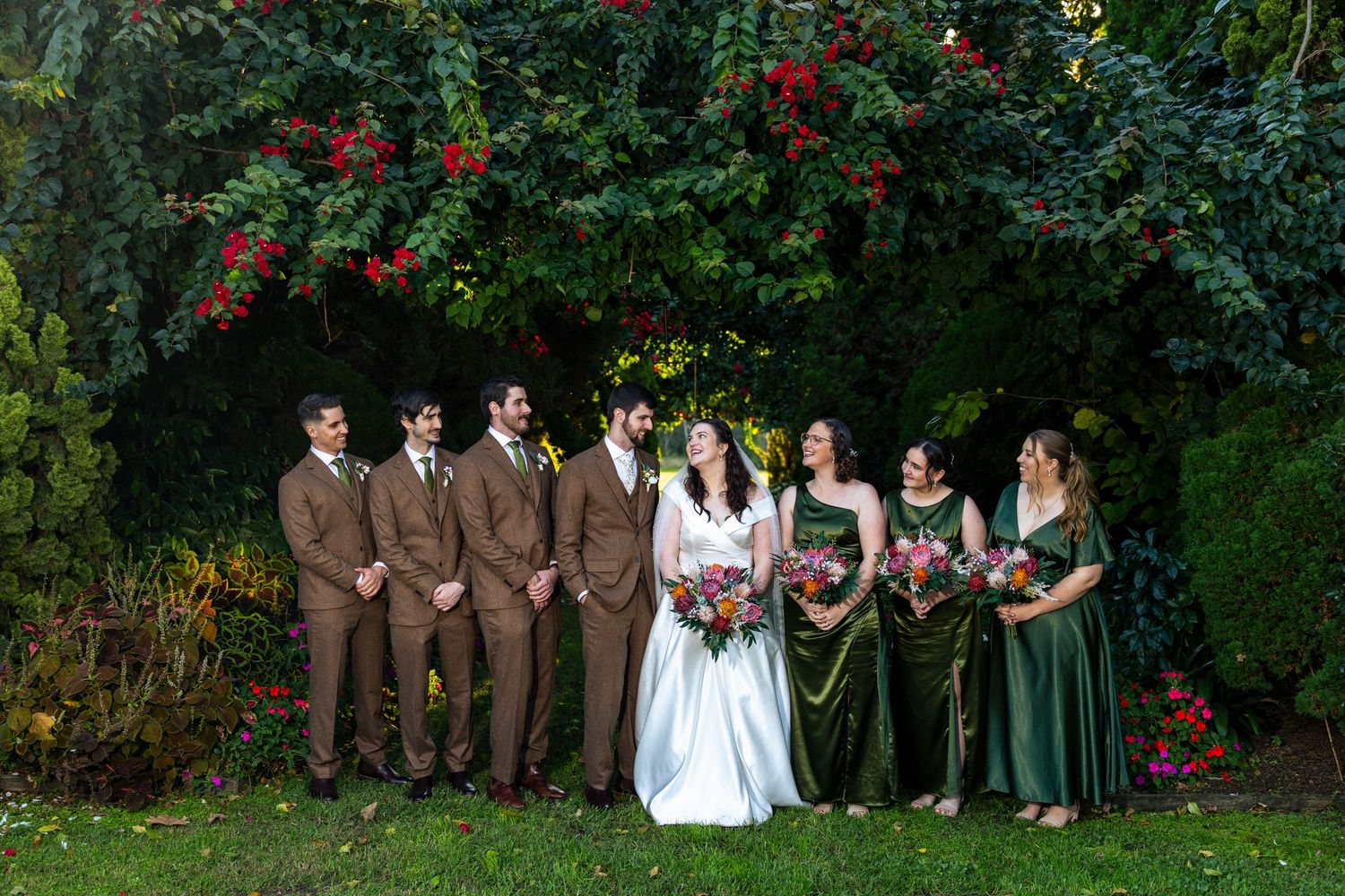 Wedding party poses outdoors under a lush hedge with red flowers at Glenleigh Estate.