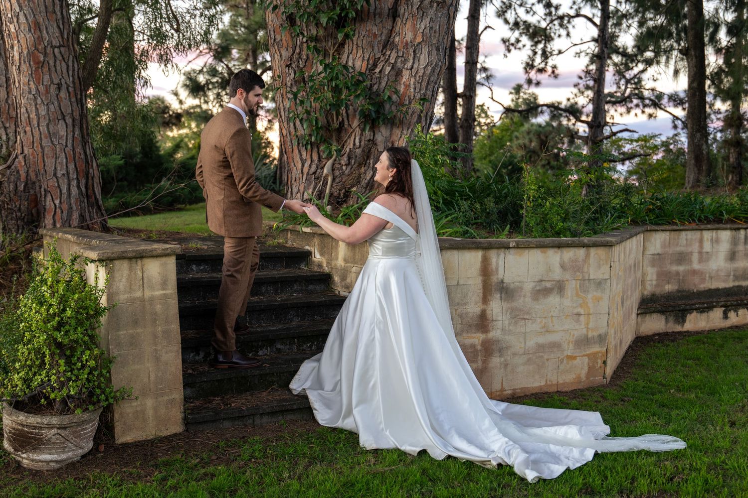 Bride in white gown reaches for groom in brown suit on stone steps surrounded by lush greenery at outdoor wedding at Glenleigh Estate.