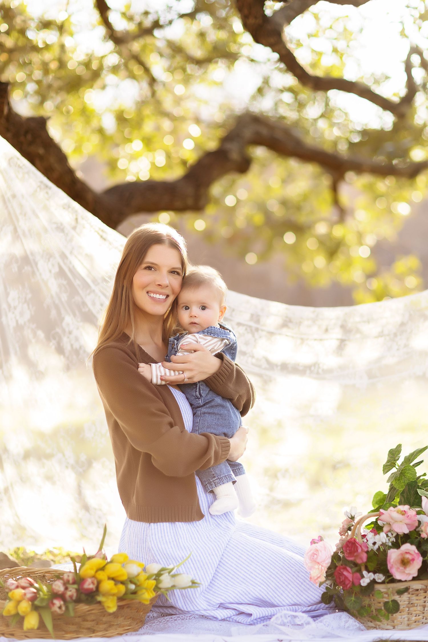 Mother and baby share tender moments on white blanket outdoors with spring flowers in golden sunlight.