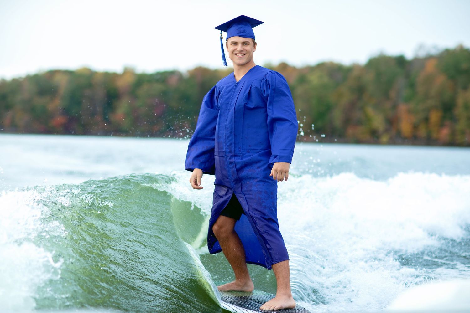 A graduate in blue cap and gown wakesurfs on a lake surrounded by fall foliage.