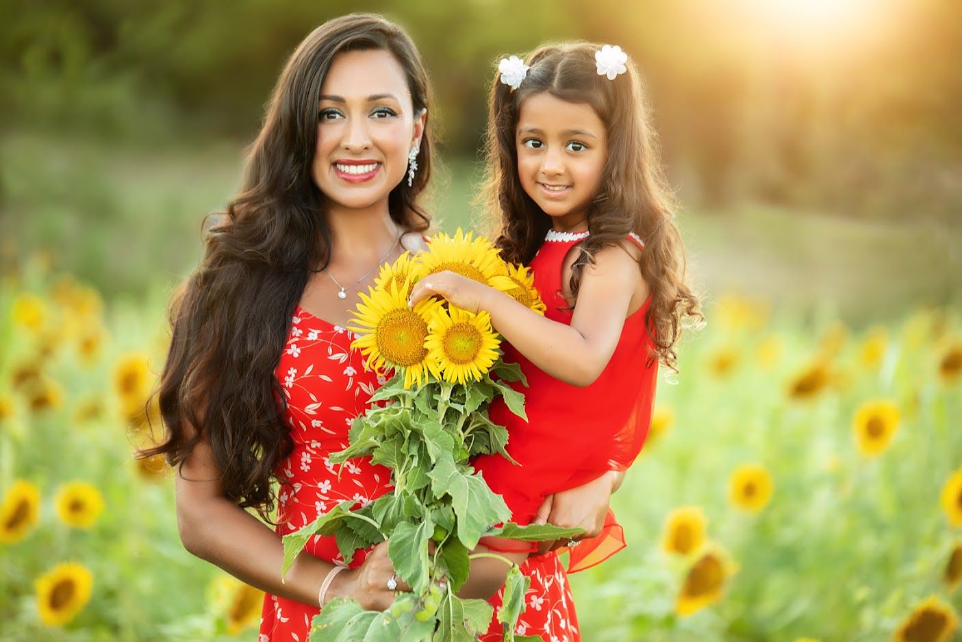 Mother and daughter in matching red dresses pose with sunflowers in a sunny field.