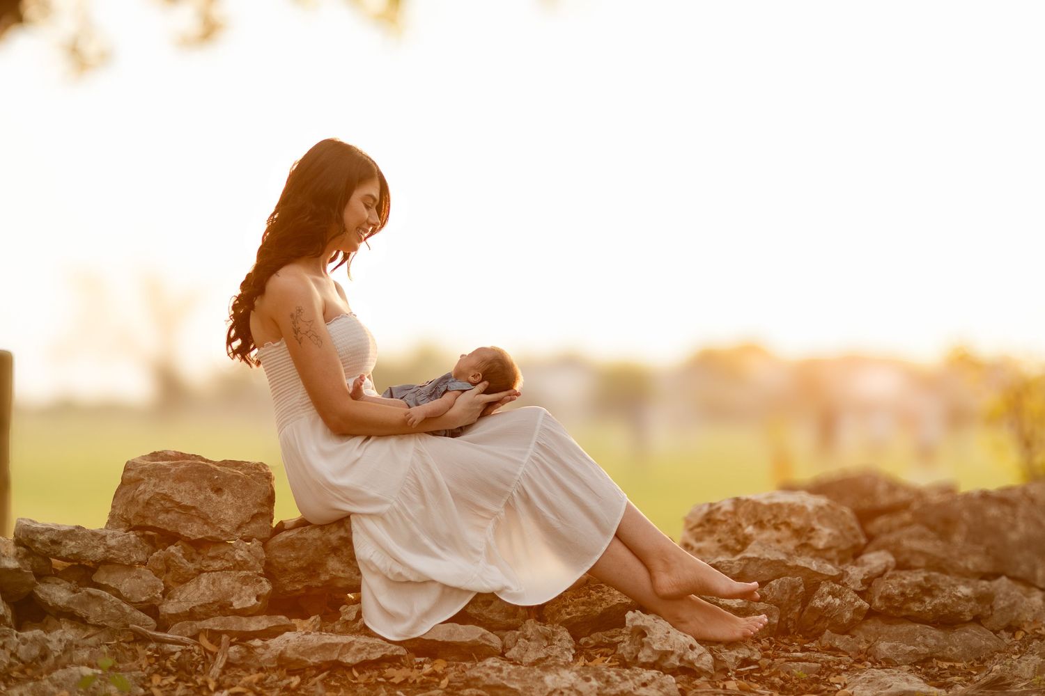 A woman in white dress sits on stone wall holding baby during golden hour.