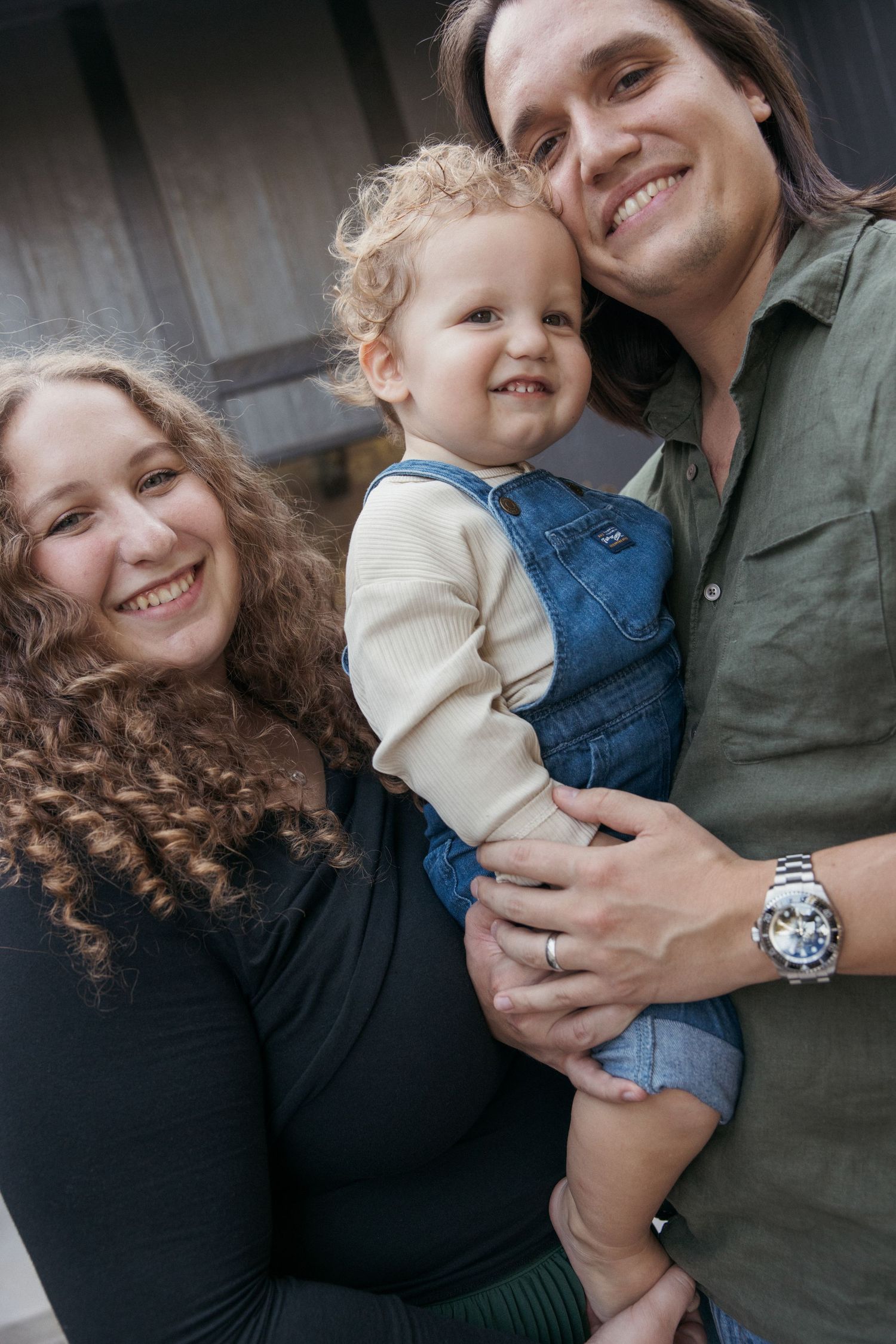 A close-up portrait of a family smiling together, with a toddler in denim overalls held between two adults.