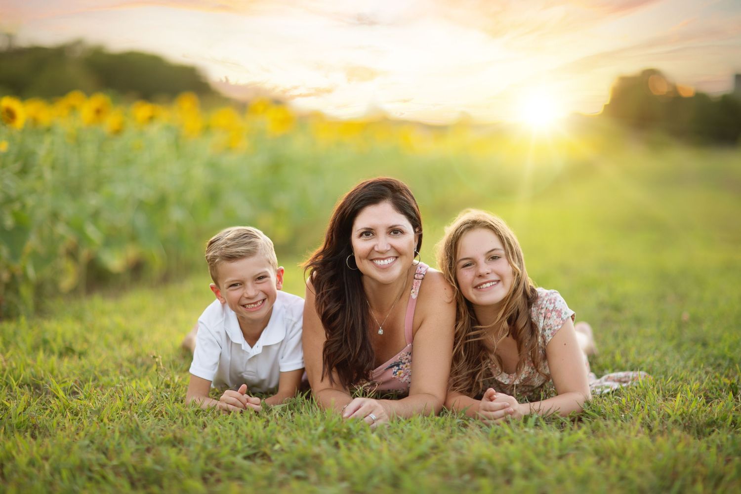 Three people laying in grass field at sunset with sunflowers in background.