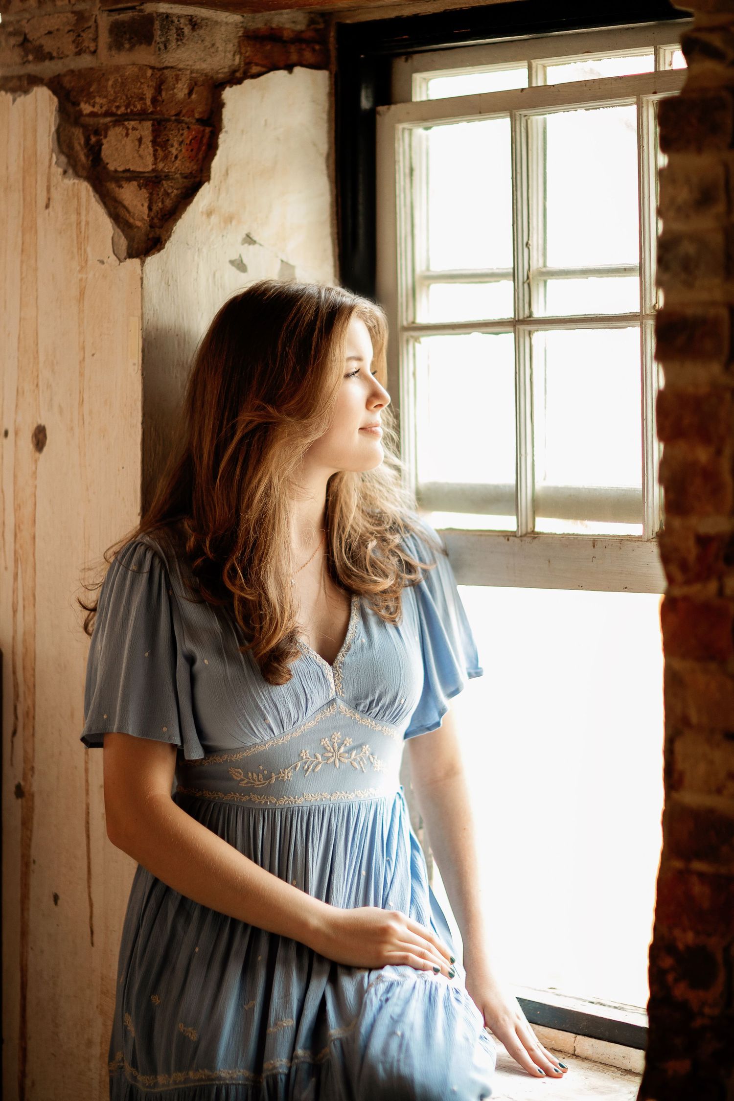 A woman in a light blue vintage dress poses by a rustic window with natural lighting.