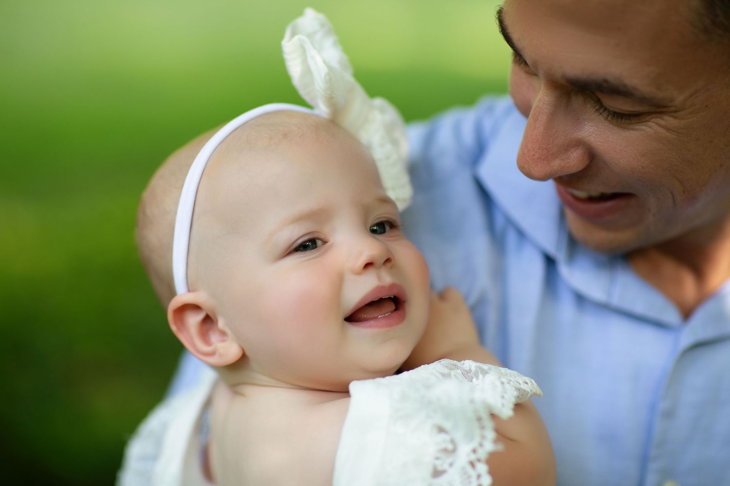 A baby wearing a white headband smiles during an outdoor portrait session.
